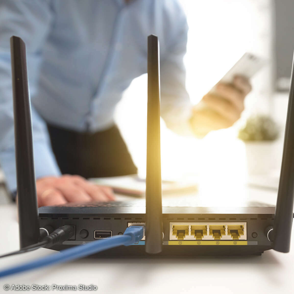 Router auf einem Schreibtisch. Im Hintergrund nutzt ein Mann Smartpohone und Laptop